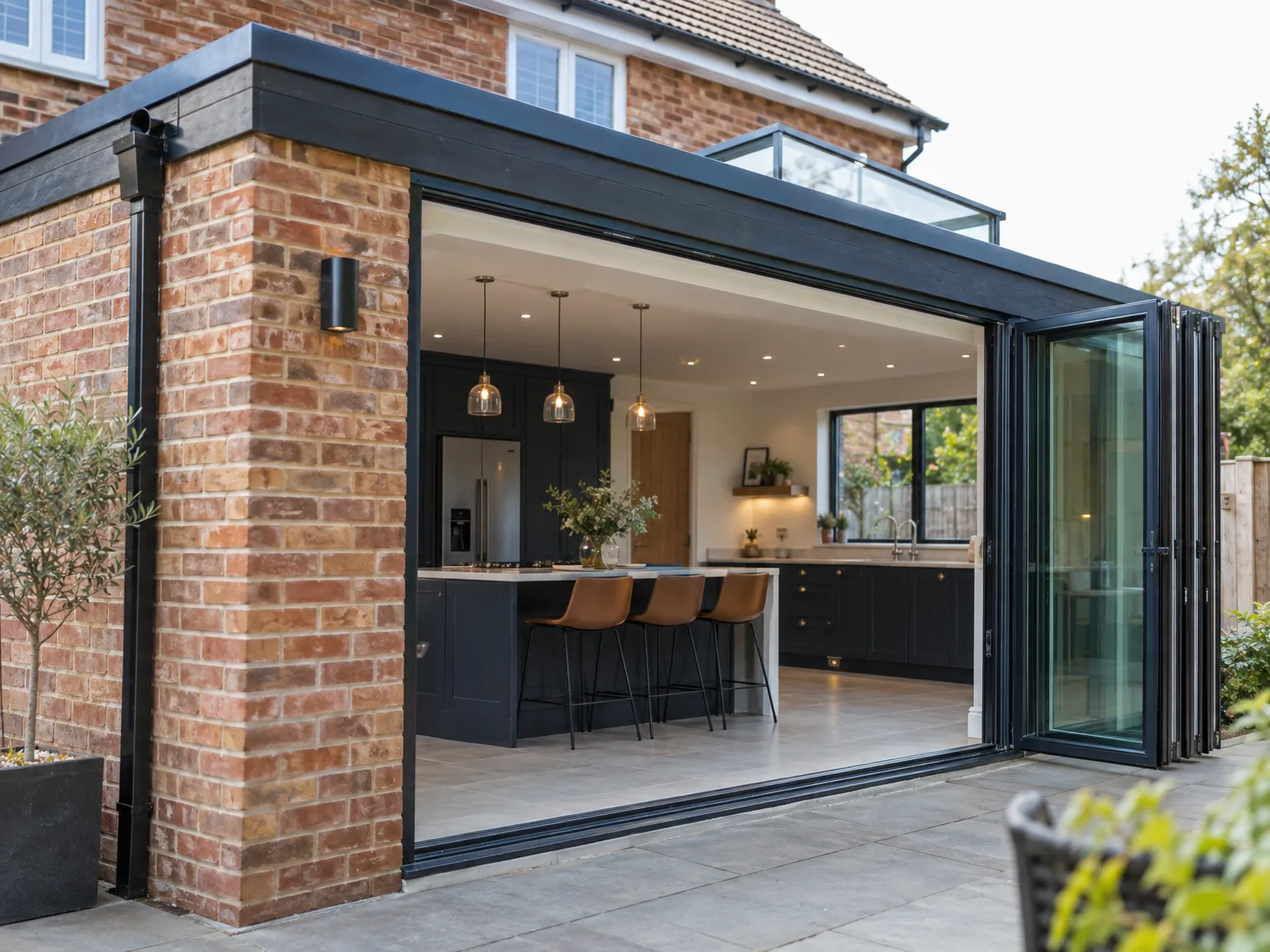 Black bifold doors opening to modern kitchen extension with island breakfast bar, brown stools, and pendant lights